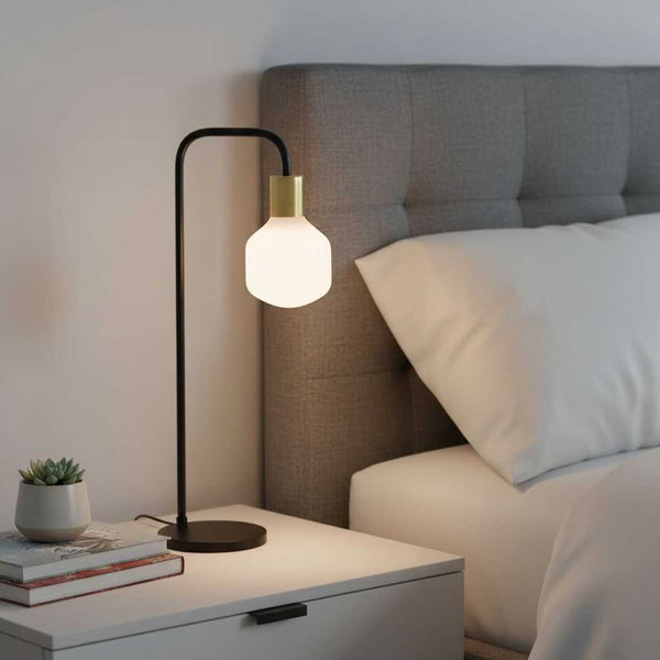 Ampoule opale allumée dans une lampe de chevet en métal noir et laiton, posée sur une table de nuit près d'un lit