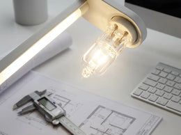 Capsule halogène allumée dans la tête d'une lampe de bureau, au-dessus de plans d'architecte et d'un pied à coulisse