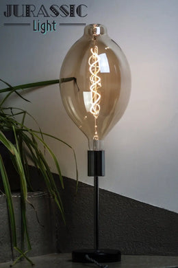 Ampoule ovale Peggy à filament LED en spirale, allumée sur un pied de lampe noir près d'une plante verte